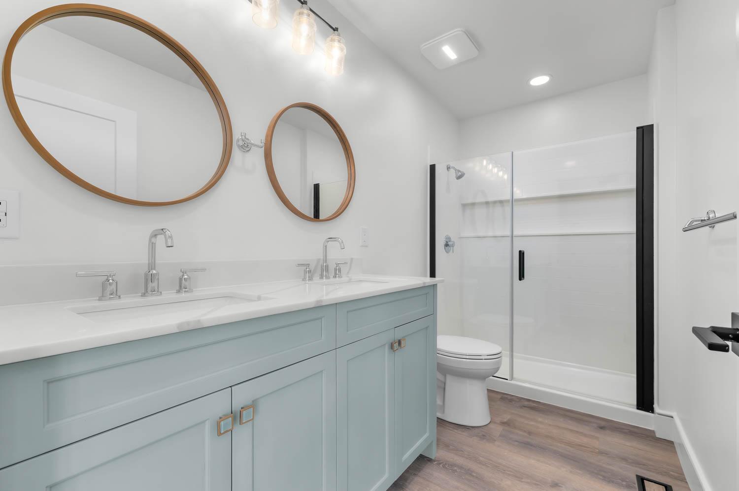 Primary bathroom remodel with round wood-framed mirrors, blue-gray double vanity, and a glass-enclosed shower