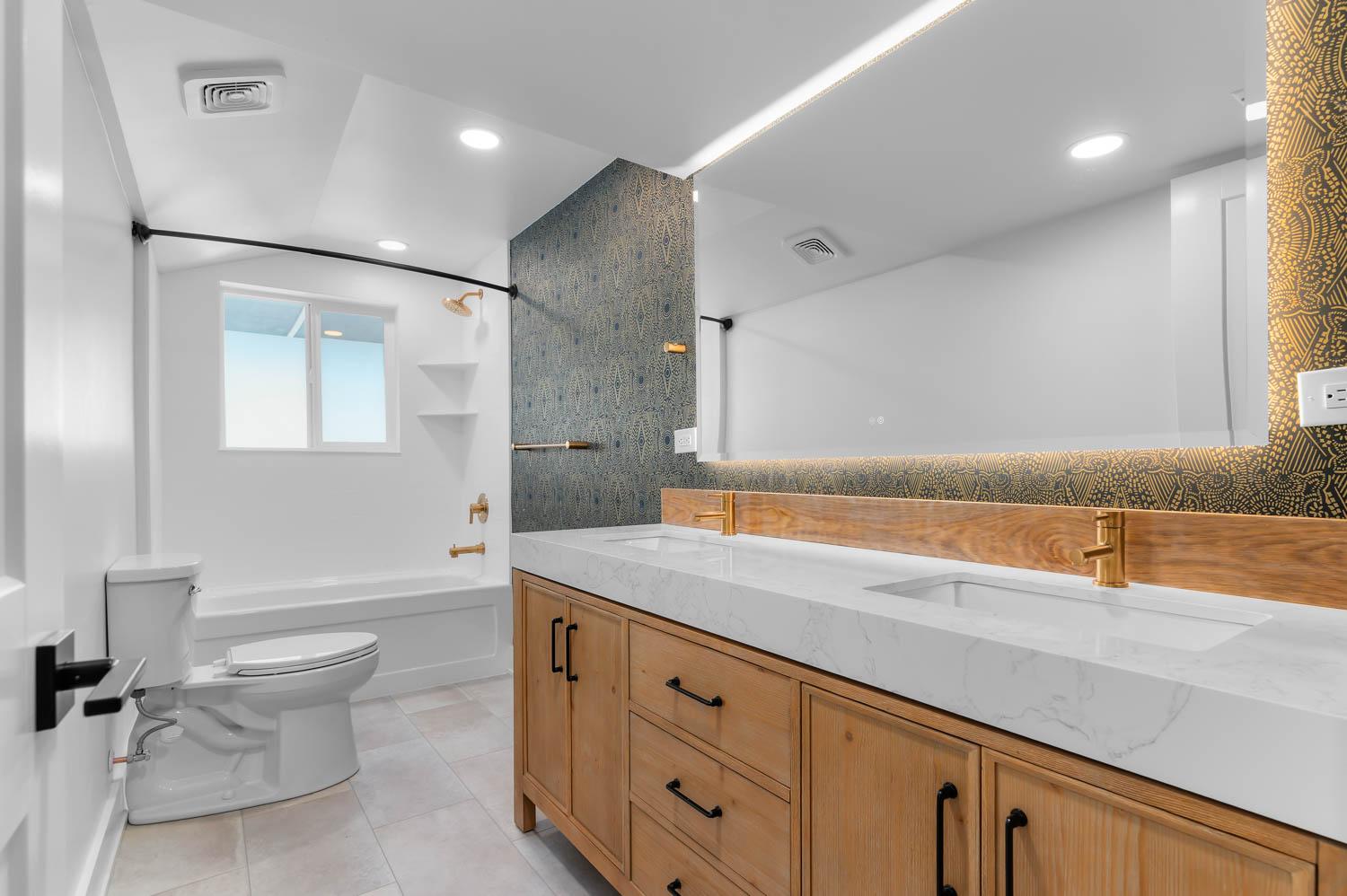 Bathroom remodel with patterned accent wallpaper, live-edge wood backsplash, and brass double-vanity fixtures